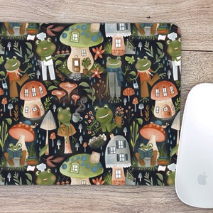 May include: A black mouse pad with a repeating pattern of green frogs, mushrooms, and houses. The frogs are wearing clothes and holding objects like books, cups, and plants. The mushrooms have red and white tops and brown stems. The houses are small and have windows and doors.