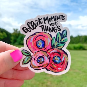 May include: A white sticker with a floral design and the text "Collect Moments Not Things". The flowers are pink, red, and white with gold accents. The leaves are green.
