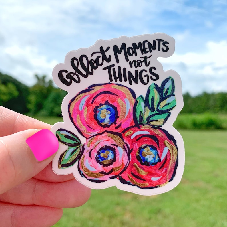 May include: A white sticker with a floral design and the text "Collect Moments Not Things". The flowers are pink, red, and white with gold accents. The leaves are green.