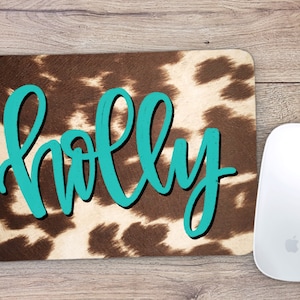 May include: A rectangular mousepad with a cowhide pattern and the name "HOLLY" in turquoise script. A white wireless mouse is positioned to the right of the mousepad. The mousepad is on a wooden surface.