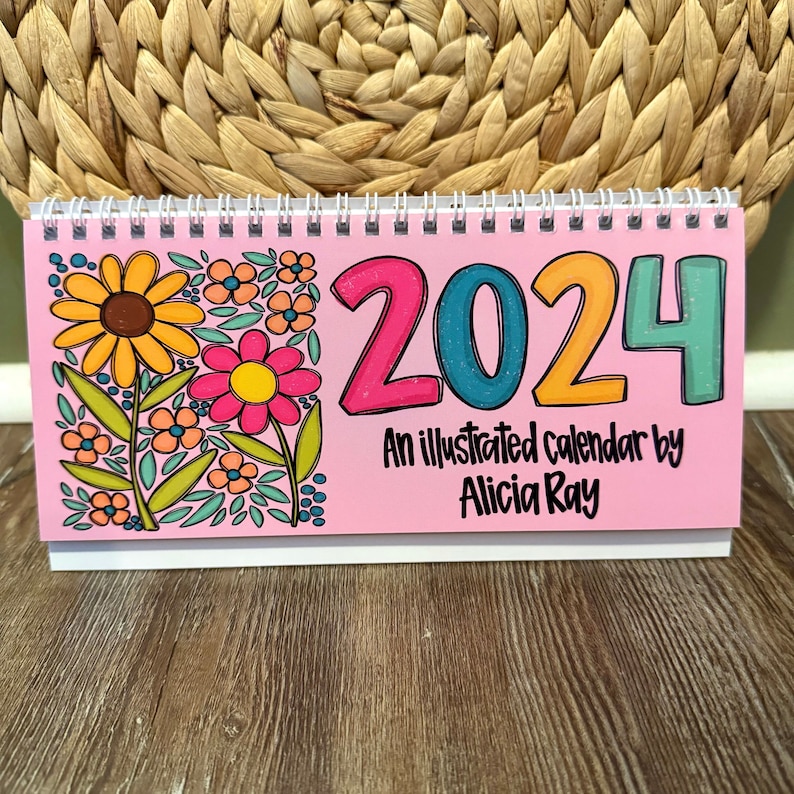 May include: A pink desk calendar for 2024 with a floral design. The calendar features the number 2024 in bright colors and the text "An illustrated calendar by Alicia Ray". The calendar is spiral-bound and sits on a white base.
