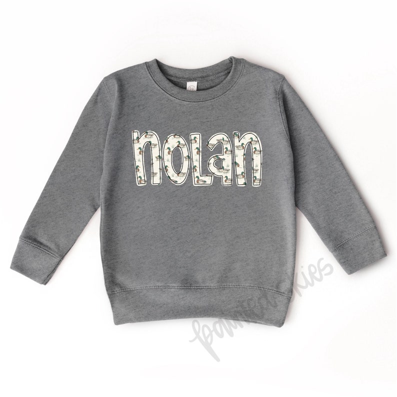 May include: Gray sweatshirt with a white outline of the name "Nolan" with a pattern of ducks inside the letters.