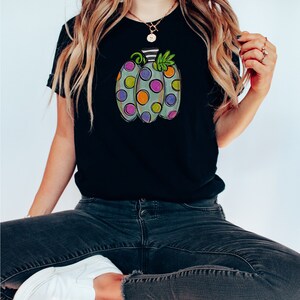 K&ouml;nnte beinhalten: Schwarzes T-Shirt mit einem bunten Cartoon-K&uuml;rbis-Design. Der K&uuml;rbis ist hellgr&uuml;n mit gro&szlig;en, farbenfrohen Tupfen in Lila, Orange und Gr&uuml;n. Der Stiel ist schwarz-wei&szlig; gestreift, mit gr&uuml;nen Bl&auml;ttern.