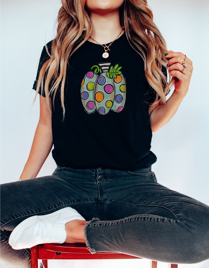 K&ouml;nnte beinhalten: Schwarzes T-Shirt mit einem bunten Cartoon-K&uuml;rbis-Design. Der K&uuml;rbis ist hellgr&uuml;n mit gro&szlig;en, farbenfrohen Tupfen in Lila, Orange und Gr&uuml;n. Der Stiel ist schwarz-wei&szlig; gestreift, mit gr&uuml;nen Bl&auml;ttern.