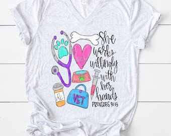 May include: Light gray v-neck t-shirt featuring a colorful veterinary-themed graphic. The design includes a stethoscope, paw print, heart, bone, pet bowl, syringe, prescription bottle, and a blue bag with the word "VET". Text reads "She works willingly with her hands Proverbs 31:13."