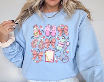 May include: Light blue sweatshirt with a graphic print of medical-themed illustrations, including a stethoscope, pills, a clipboard, a pair of clogs, a bottle of oxygen, and a pair of lungs. The graphic is in pink, blue, and white.