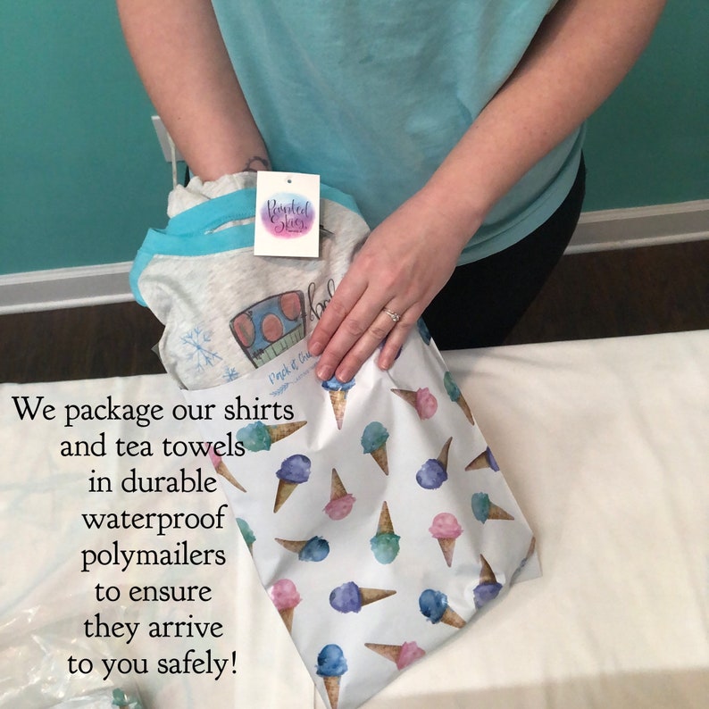 Pode incluir: Uma camisa cinza com acabamento azul e uma etiqueta "Painted Skies" est&aacute; sendo embalada em um polymailer branco. O polymailer tem um padr&atilde;o de cones de sorvete em aquarela em azul, rosa e verde.