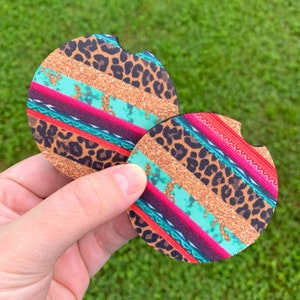 May include: Two round car coasters with a colorful design. The coasters feature a pattern of leopard print, gold glitter, turquoise, and red stripes. The coasters are held in a hand against a green background.