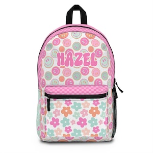 May include: A pink backpack with a black handle and zipper. The backpack features a pattern of smiley faces in pastel colors and the name "HAZEL" in pink. The front pocket has a pink checkered design, and the bottom has a floral pattern.