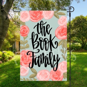 May include: A garden flag with a light blue background and pink floral design. The text "the book family" is written in black cursive lettering.