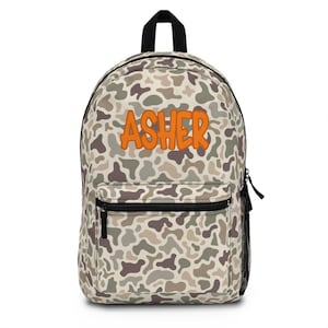 May include: A camouflage-patterned backpack with the name "ASHER" in orange block letters. The backpack has a front zippered pocket, a black handle, and black shoulder straps. The camouflage design features shades of brown, gray, and beige.