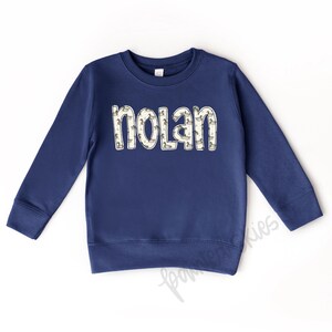 May include: A navy blue long-sleeved sweatshirt with the name "Nolan" printed on the front in white letters outlined with a pattern of small ducks.