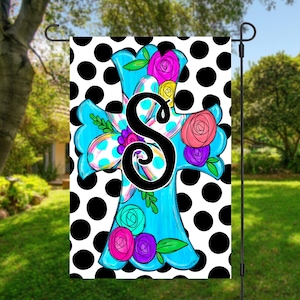 May include: A blue cross with white polka dots and colorful flowers, with the letter "S" in black in the center. The cross is on a white background with black polka dots.
