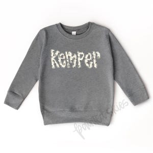 May include: A gray sweatshirt with the name "Kemper" printed on the front in white letters with a camouflage pattern.