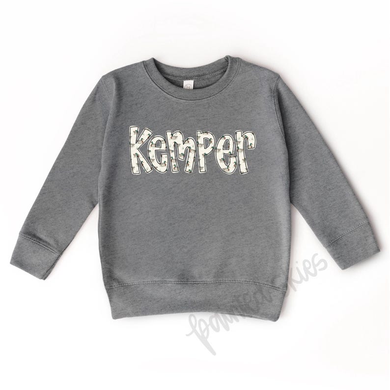 May include: A gray sweatshirt with the name "Kemper" printed on the front in white letters with a camouflage pattern.