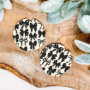 May include: Two round car coasters with a cream background and a repeating pattern of black bows. The coasters are made of a durable material and are designed to fit in most car cup holders. A stylish accessory.