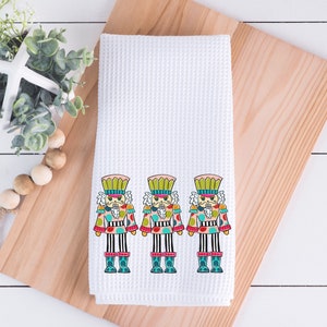May include: White waffle-weave kitchen towel featuring three colorful nutcracker soldier designs. The nutcrackers have pink, green, blue, and red accents. The towel is displayed on a wooden cutting board.