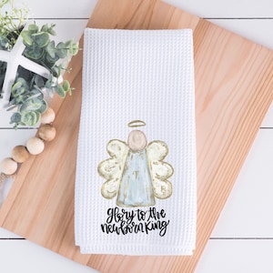 Pode incluir: Toalha de cozinha branca com tecido waffle, com um desenho de anjo em aquarela com detalhes dourados e azul claro. O texto "Glory to the newborn King" est&aacute; impresso em preto. A toalha &eacute; exibida em uma t&aacute;bua de corte de madeira.