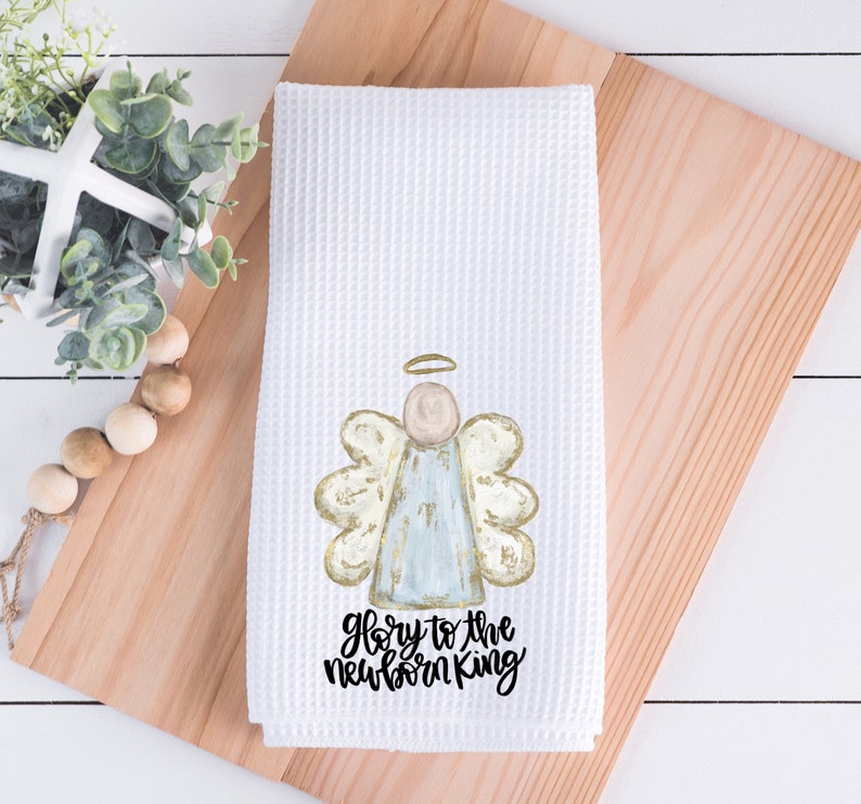 Pode incluir: Toalha de cozinha branca com tecido waffle, com um desenho de anjo em aquarela com detalhes dourados e azul claro. O texto "Glory to the newborn King" est&aacute; impresso em preto. A toalha &eacute; exibida em uma t&aacute;bua de corte de madeira.