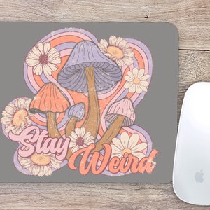 May include: A gray mouse pad with a retro-style design featuring colorful mushrooms and flowers. The text "Stay Weird" is written in a groovy font.