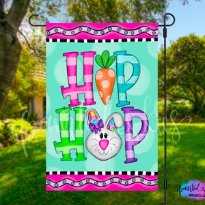 May include: A colorful garden flag with the word "HOP" in large, multi-colored letters. A cartoon bunny with a pink bow is in the center of the flag, and a carrot is behind the bunny. The flag has a pink, white, and blue border.