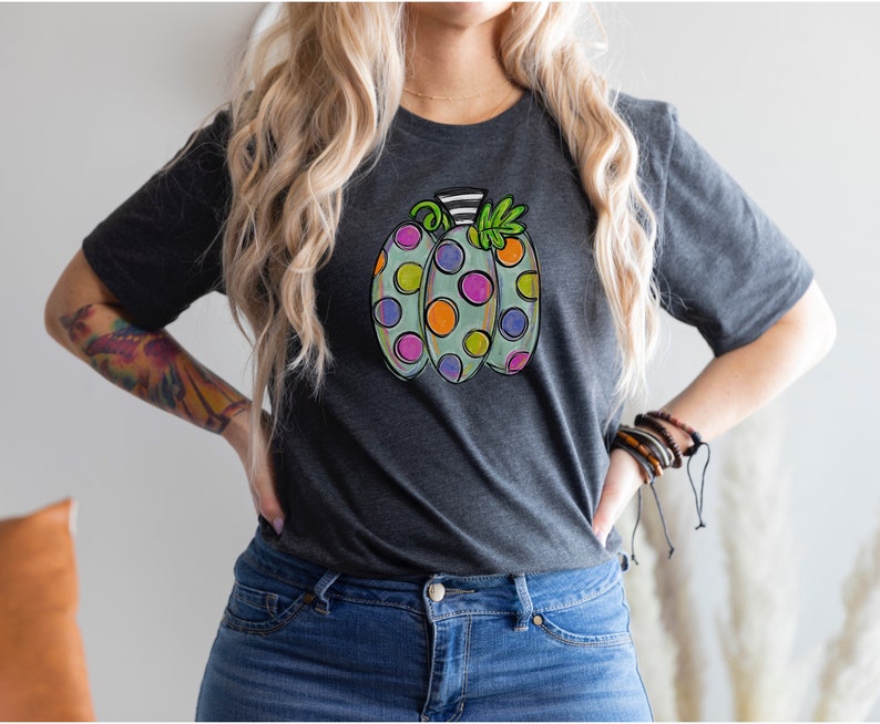 K&ouml;nnte beinhalten: Dunkelgraues T-Shirt mit einem bunten K&uuml;rbisdesign. Der K&uuml;rbis ist hellgr&uuml;n mit gro&szlig;en, bunten Tupfen in Lila, Orange und Gelb. Der Stiel ist schwarz-wei&szlig; gestreift. Ein lustiges und festliches, herbstliches Grafik-T-Shirt.