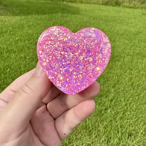 May include: A heart-shaped object with a smooth, glossy finish, featuring a vibrant pink base color embedded with a dense mixture of multi-colored, reflective glitter particles that create a sparkling effect across the entire surface.