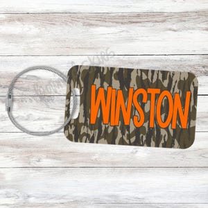 May include: A rectangular luggage tag with a camouflage pattern in shades of brown and green. The name "WINSTON" is printed in bold orange letters. A silver-colored metal loop is attached for securing the tag.