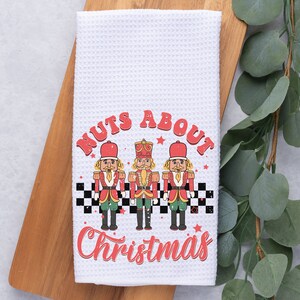 May include: White kitchen towel with a red and black design featuring three nutcracker figures and the text "Nuts About Christmas".
