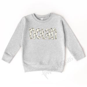 May include: A gray sweatshirt with a white outline of the name "Nolan" printed on the front. The outline is filled with a pattern of small ducks.