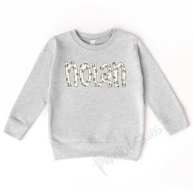 May include: A gray sweatshirt with a white outline of the name "Nolan" printed on the front. The outline is filled with a pattern of small ducks.