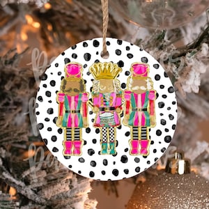 May include: A round Christmas ornament with a white background and black polka dots. Three nutcracker figures are featured, one playing a drum, with pink, green, and gold accents.