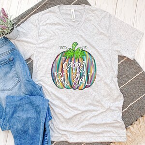 May include: A light gray v-neck t-shirt featuring a colorful pumpkin design with the text "Happy Fall Y'all". The pumpkin is painted with vertical strokes of various colors. The shirt is paired with blue jeans.