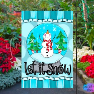 May include: A blue and white striped garden flag with a snow globe design. The snow globe features a snowman with a red scarf and hat, and two small green trees. The text "Let it Snow" is printed in black below the snow globe.