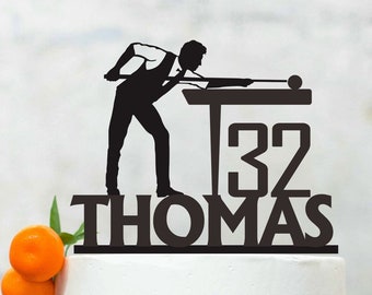 May include: A white cake with orange decorations and a black silhouette of a person playing pool with the number 32 and the name Thomas.