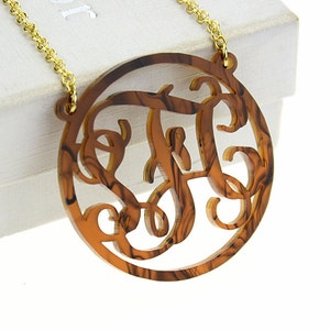 May include: A gold chain necklace with a brown marbled round pendant featuring a cut-out monogram. The pendant has a swirl pattern.