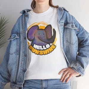 May include: White t-shirt featuring a cartoon elephant with purple ears and a blue body, set against a yellow sunburst. The text "HELLO SUNSHINE" is written in yellow. The model is wearing a denim jacket.