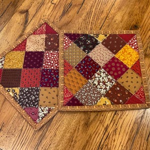 May include: Two square quilted potholders featuring a patchwork design of various fabric squares in shades of red, yellow, brown, and cream. Each potholder is finished with a decorative tan border featuring a repeating floral pattern.
