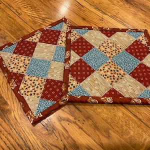 May include: Two quilted pot holders with a patchwork design in shades of red, brown, beige, and blue. The pot holders are made of fabric with different patterns, including floral, geometric, and paisley.