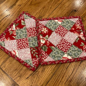May include: Two square quilted pot holders featuring a patchwork design of red, white, and green fabric squares. The patterns include floral motifs, holly leaves, and small white berries on red and green backgrounds, all finished with a red border.