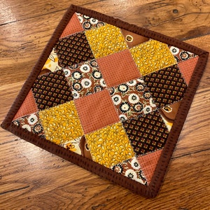 May include: A brown and yellow patchwork quilted hot pad with a brown border. The hot pad is made of different fabrics with floral, leaf, and geometric patterns.