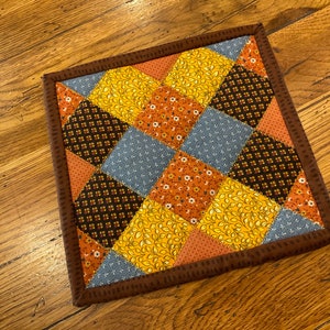 May include: A brown and orange patchwork quilt square with a brown border. The quilt square is made of different fabrics with floral and leaf patterns.