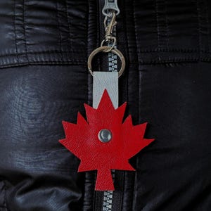 May include: A red maple leaf keychain with a silver-colored metal rivet in the center. The keychain is attached to a silver-colored metal ring and a light gray leather strap. The background is a black quilted jacket.