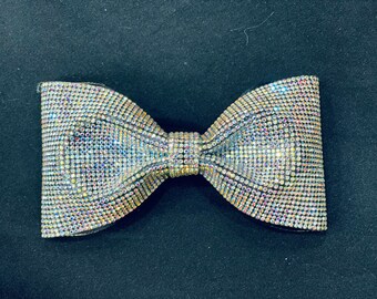 May include: A large, iridescent bow made of rhinestones. The bow is a light silver color with a slight rainbow effect.