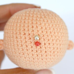 Peut inclure: Une t&ecirc;te de poup&eacute;e en crochet de couleur p&ecirc;che. La t&ecirc;te de poup&eacute;e a deux petites oreilles en crochet et trois perles sur le dessus. Les perles sont blanches, rouges et blanches.
