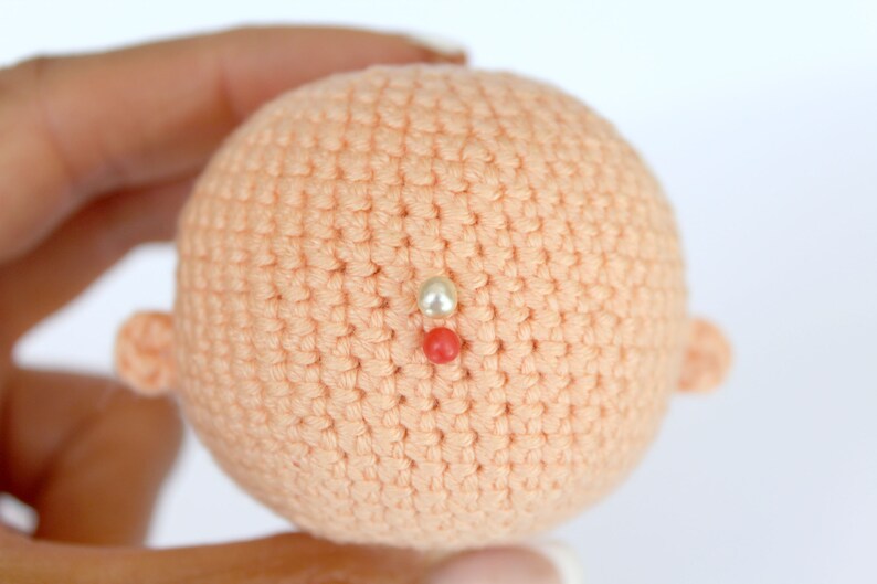 Peut inclure: Une t&ecirc;te de poup&eacute;e en crochet de couleur p&ecirc;che. La t&ecirc;te de poup&eacute;e a deux petites oreilles en crochet et trois perles sur le dessus. Les perles sont blanches, rouges et blanches.