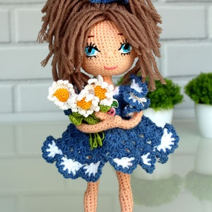 Peut inclure: Une poup&eacute;e en crochet avec des cheveux bruns, des yeux bleus et une robe bleue et blanche. La poup&eacute;e tient un bouquet de marguerites blanches.