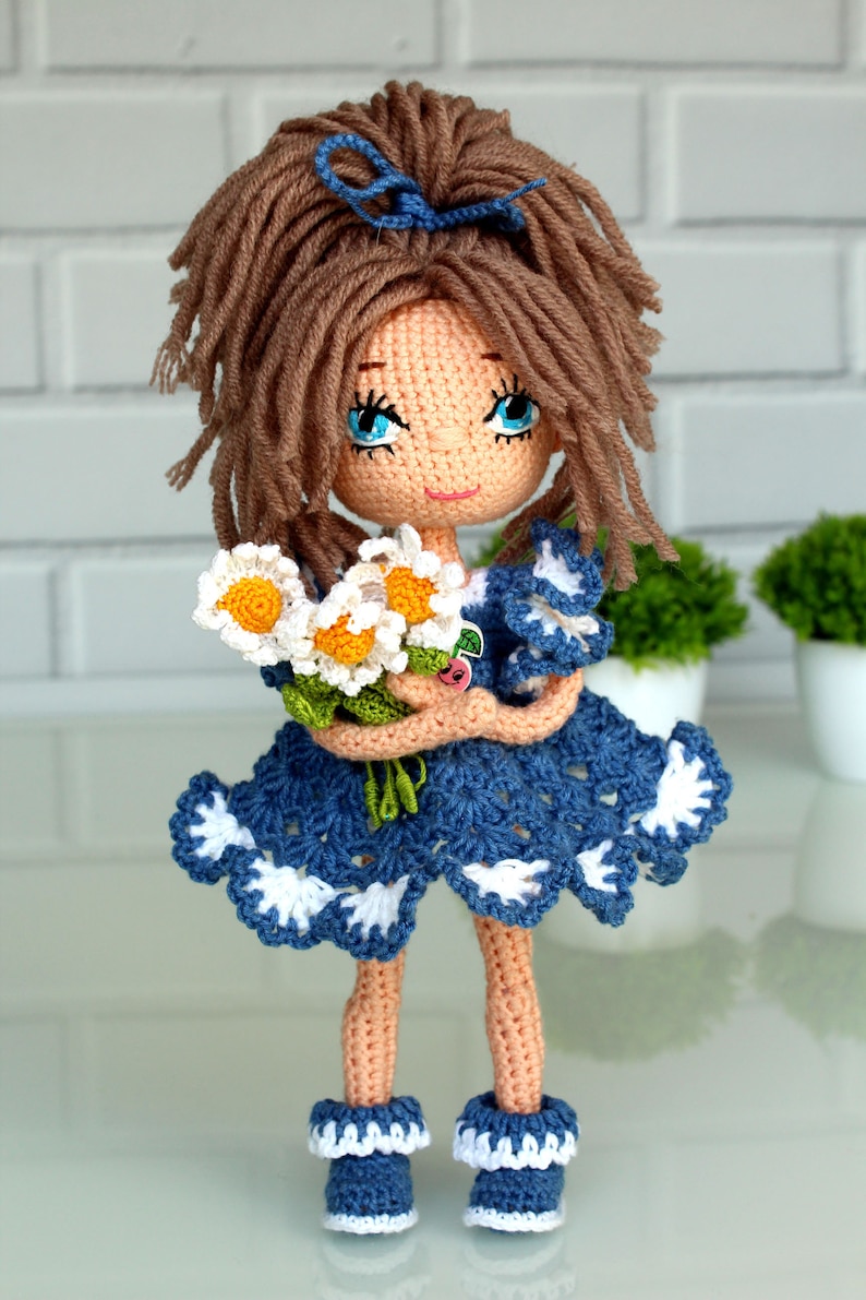 Peut inclure: Une poup&eacute;e en crochet avec des cheveux bruns, des yeux bleus et une robe bleue et blanche. La poup&eacute;e tient un bouquet de marguerites blanches.