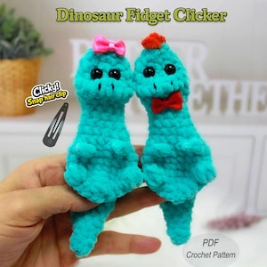 以下が含まれることがあります： ピンクのリボンと赤い蝶ネクタイとオレンジ色の房が付いた、2つのターコイズのかぎ針編みの恐竜フィジェットクリッカー。画像には、黒いスナップヘアクリップと「Dinosaur Fidget Clicker」と「Clicky! Snap hair clip」というテキストが含まれています。