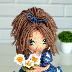 Peut inclure: Une poup&eacute;e en crochet avec des cheveux bruns, des yeux bleus et un n&oelig;ud bleu. La poup&eacute;e porte une robe bleue avec des accents blancs et tient un bouquet de marguerites blanches.